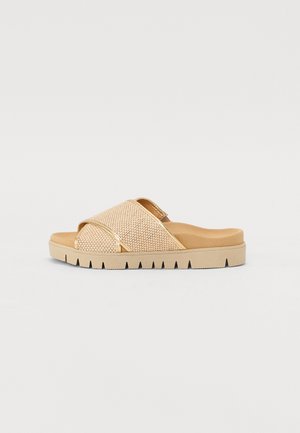 Beige slide sandal with a woven texture, cross straps, and a chunky sole featuring notched edges. Lightweight and open-toe design.
