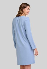 Mujer con cabello ondulado que lleva un vestido de manga larga y hasta la rodilla de color azul claro, mirando ligeramente hacia la derecha sobre un fondo gris liso.