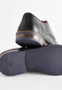 Chaussures en cuir noir avec un bout arrondi, accent de talon brun contrastant et une semelle en caoutchouc violet texturé arborant un motif hexagonal.
