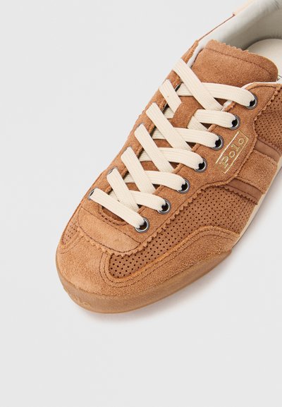 Sneaker in suede marrone con lacci beige, pannelli laterali perforati e dettaglio del logo in oro. Presenta una punta testurizzata e suola in gomma a contrasto.