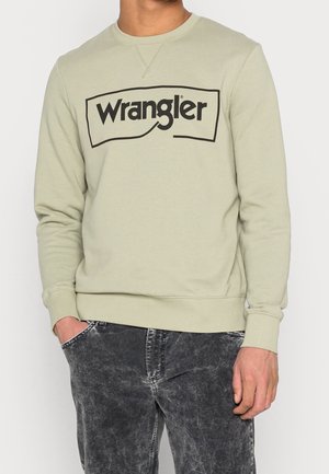Mężczyzna ubrany w jasnozieloną bluzę Wrangler z czarnym logo i wyblakłe czarne dżinsy, stojący z jedną ręką w kieszeni.