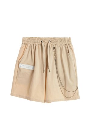 Pantalones cortos casuales beige con cintura elástica, cordón, bolsillos laterales, una etiqueta blanca en el bolsillo izquierdo y una cadena decorativa plateada en el lado derecho.
