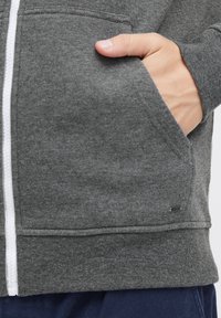 Grijze ritsswetshirt met een witte rits, een voorzak en een klein merklabel aan de zoom. Gladde stof met een zachte textuur.