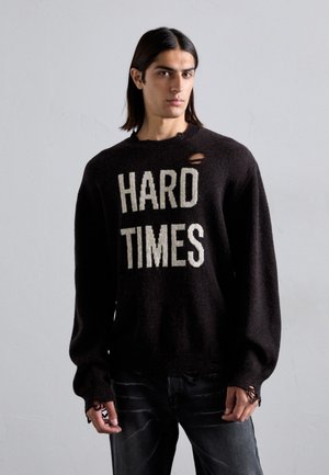 Schwarzer Strickpullover mit dem Text "HARD TIMES" in Creme. Mit Rundhalsausschnitt und ausgefranstem Detail an der Schulter. Getragen mit dunklen Jeans.