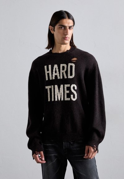 Sort strikket sweater med teksten "HARD TIMES" i creme. Har en rund halsudskæring og slidte detaljer på skulderen. Bæres med mørke jeans.