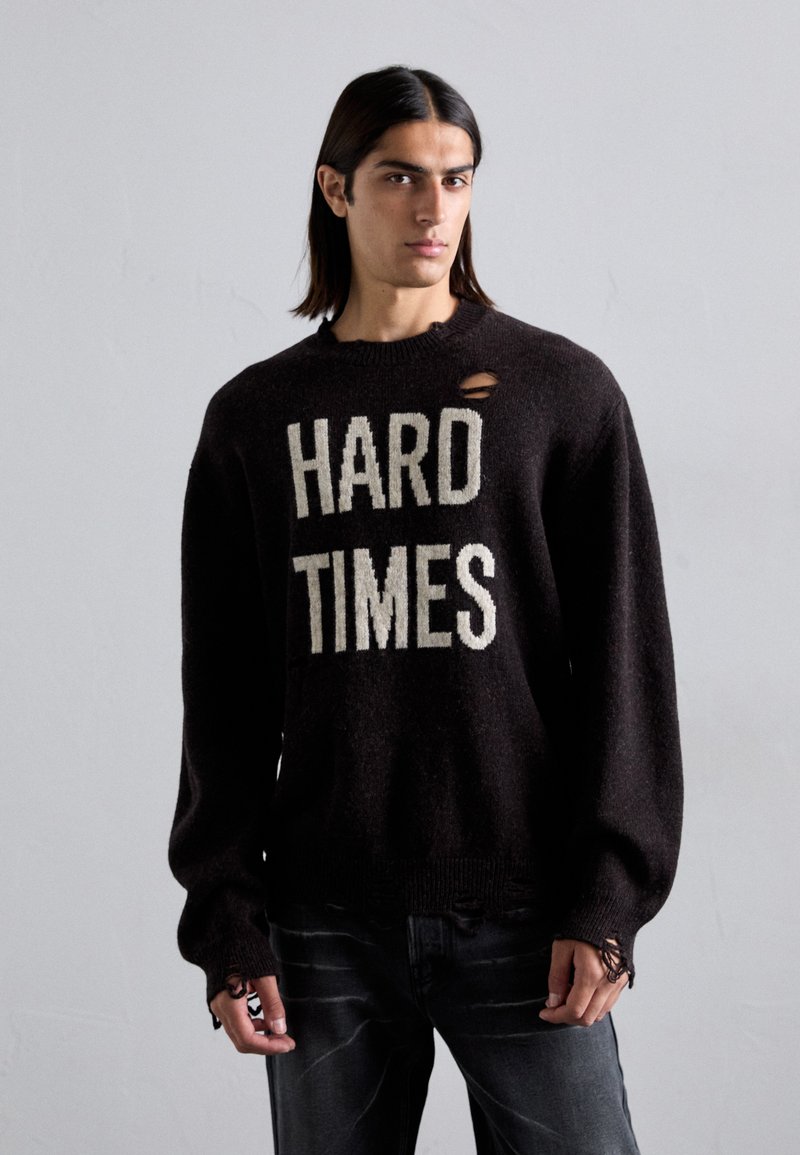 Schwarzer Strickpullover mit dem Text "HARD TIMES" in Creme. Mit Rundhalsausschnitt und ausgefranstem Detail an der Schulter. Getragen mit dunklen Jeans.