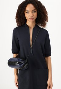 Navyblaues Kleid mit hohem Kragen und einem Reißverschluss vorne, ausgestattet mit zwei Fronttaschen. Kleine Handtasche aus navyblauem und schwarzem Leder mit einem geknoteten Design.