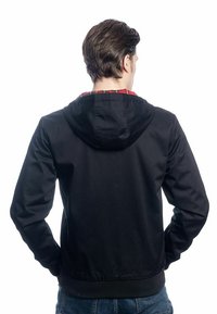 Veste à capuche noire avec une texture lisse, des poignets et un ourlet élastiques, présentant une doublure à carreaux rouges visible au niveau de la capuche.