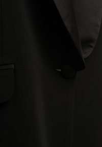 Blazer nero con una texture liscia, revers a punta e chiusura con bottone rotondo vicino al colletto. Design minimalista con tasca laterale.