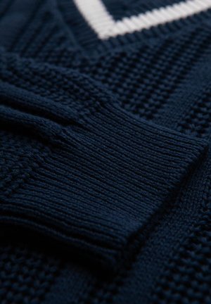 Granatowy sweter z dzianiny z wypukłymi wzorami i białym dekoltem w serek. Posiada ściągacze przy rękawach oraz różnorodne wzory ściegów na całej powierzchni.