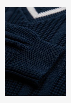 Granatowy sweter z dzianiny z wypukłymi wzorami i białym dekoltem w serek. Posiada ściągacze przy rękawach oraz różnorodne wzory ściegów na całej powierzchni.