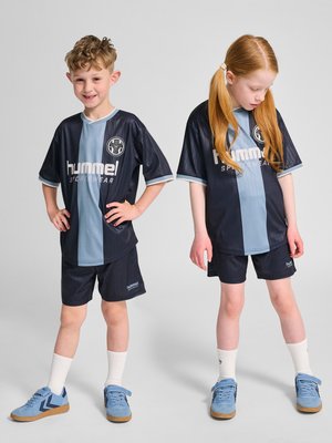 To børn står side om side iført matchende sort og lyseblåt sportstøj med hvide sokker og blå sneakers mod en ensfarvet baggrund.