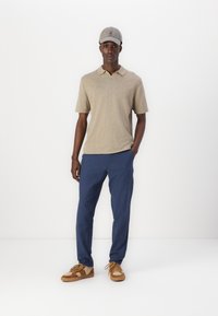 Camisa polo beige, pantalones azules y zapatillas marrones con detalles de ante. Los accesorios incluyen una gorra gris. Un atuendo sencillo y casual.