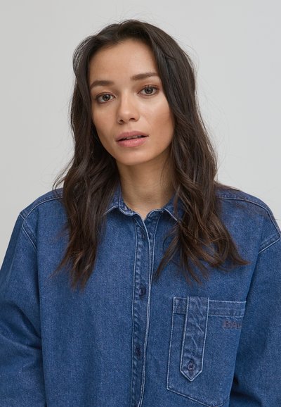 Blå denimskjorta med spetsig krage, långa ärmar och bröstficka med diskreta logodetaljer. Mjuk textur och avslappnad passform.