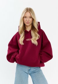 Felpa oversize bordeaux con tasca a marsupio e polsini a coste. Presenta un logo ricamato sul davanti, abbinata a jeans azzurri.