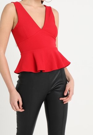 Femme portant un haut péplum rouge sans manches à décolleté plongeant, associé à un pantalon noir ajusté taille haute, sur fond blanc.