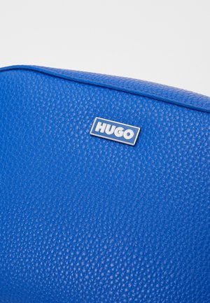 Mała niebieska skórzana torebka na ramię z regulowanym paskiem i srebrnym logo "HUGO" z przodu na białym tle.