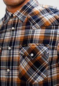 Brun och marinblå rutiga flanellskjorta med knäppkrage, två bröstfickor och vita knappar. Mjuk textur med ett rutigt mönster.