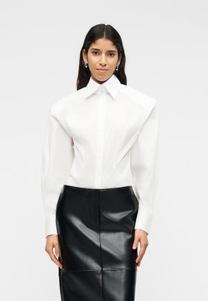 Kvinde med langt mørkt hår iført hvid struktureret bluse med overdrevne skuldre og sort lædernederdel, stående foran en ensfarvet baggrund.