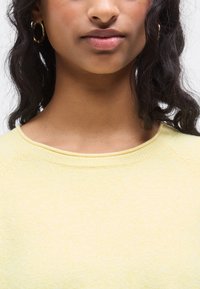 Pull jaune clair texturé avec un large col rond. Comprend des manches raglan et une finition en maille douce et confortable. Variation de couleur subtile visible.