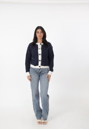 Cardigan lavorato a maglia blu navy con dettagli crema, caratterizzato da una parte frontale texturizzata, grandi bottoni e polsini a costine, abbinato a jeans blu chiaro.