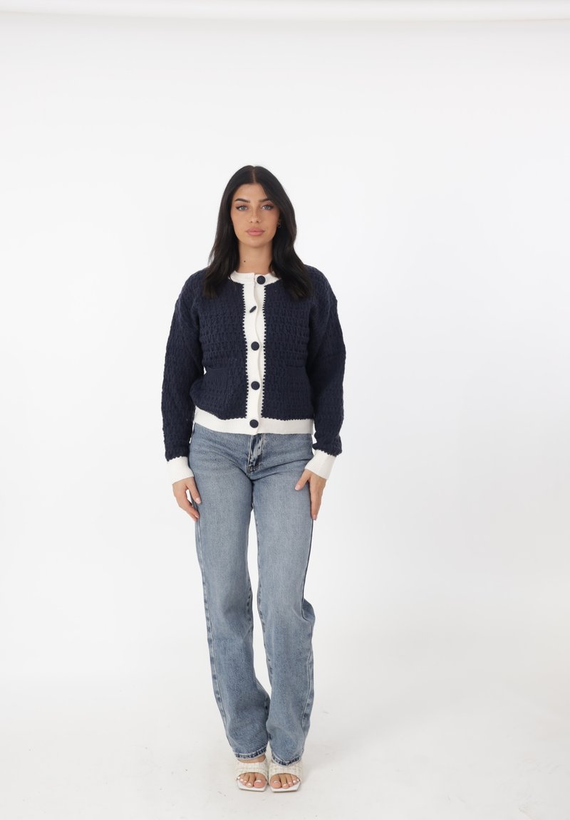 Cardigan lavorato a maglia blu navy con dettagli crema, caratterizzato da una parte frontale texturizzata, grandi bottoni e polsini a costine, abbinato a jeans blu chiaro.