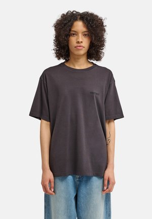 Młoda osoba o kręconych włosach, ubrana w luźny ciemnoszary t-shirt i jasnoniebieskie dżinsy, stojąca na jednolitym jasnym tle.
