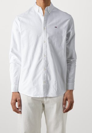 Hombre con una camisa blanca de manga larga abotonada con un pequeño logo rojo, blanco y azul en el pecho, combinado con pantalones color crema.
