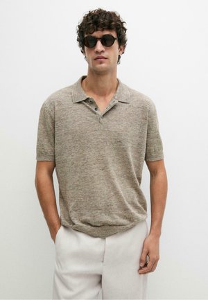 Man draagt ronde zwarte zonnebril, beige korte mouw gebreide polo en lichte broek tegen een effen witte achtergrond.