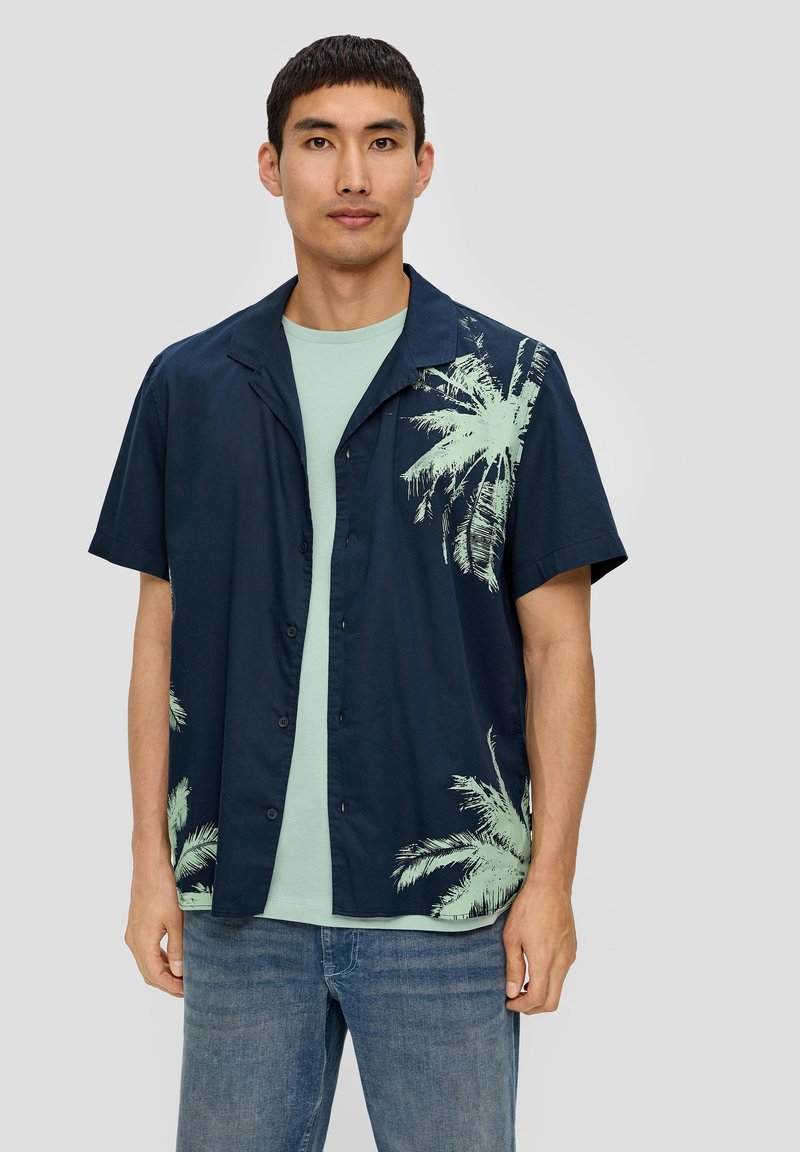 Marineblauwe short sleeve shirt met mintgroene palmboomgraphics aan één kant, gecombineerd met een lichtgroene T-shirt eronder en blauwe jeans.