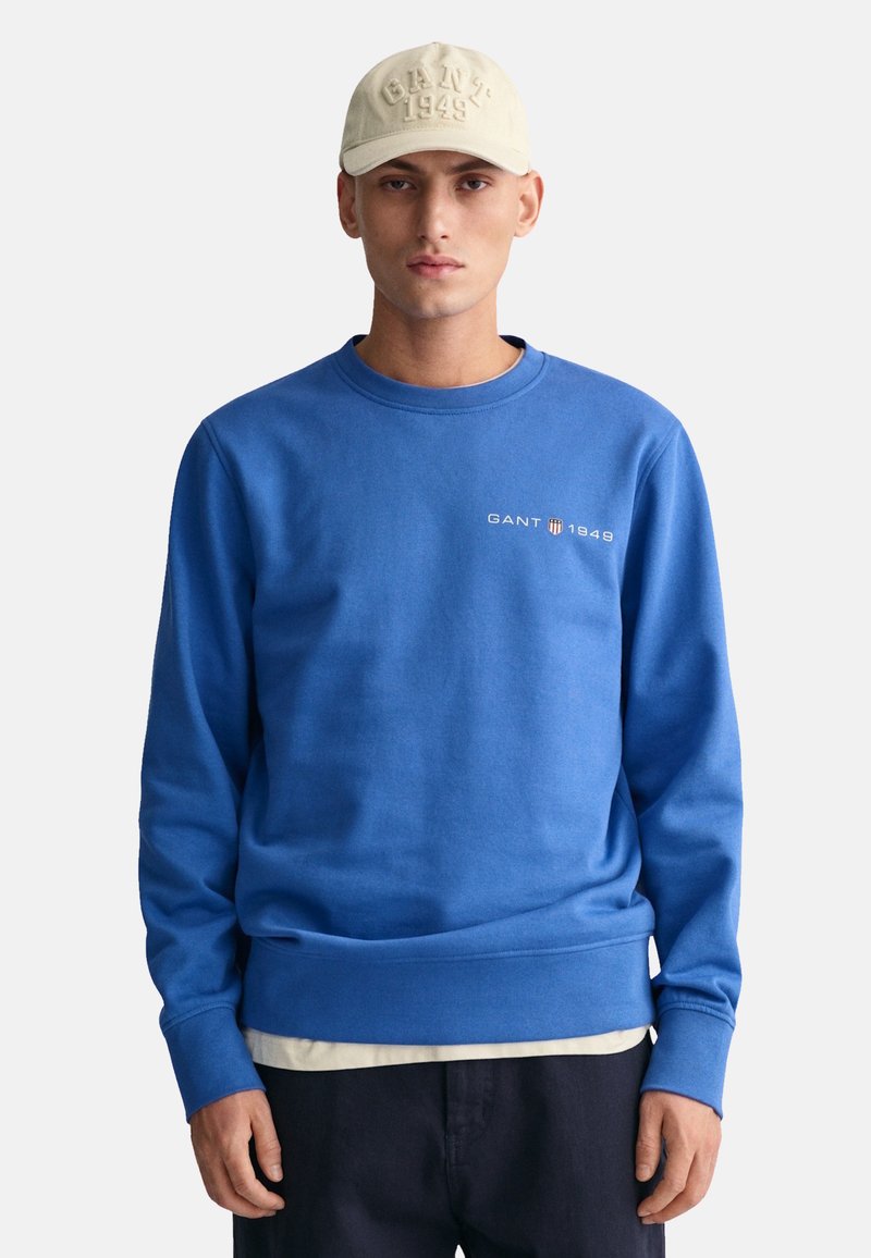 Felpa blu realizzata in misto cotone, con polsini e orlo a coste, con "GANT 1949" ricamato sul petto. Abbinata a un berretto beige.