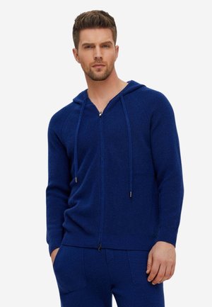 Mann trägt einen blauen Kapuzenpullover mit Reißverschluss und Kordeln sowie passende blaue Hose, steht mit teilweise in den Taschen steckenden Händen.