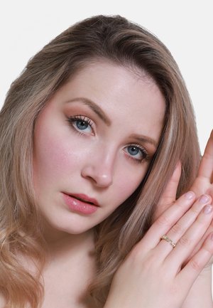 Frau mit langen, welligen blonden Haaren, blauen Augen und natürlichem Make-up. Sie trägt einen dünnen goldenen Ring an ihrer linken Hand, der in der Nähe ihres Gesichts positioniert ist.