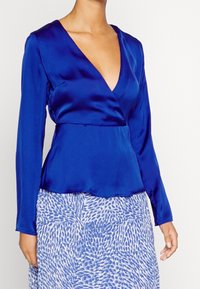 Top avvolgente in raso blu con maniche lunghe e scollatura a V profonda, caratterizzato da una vita aderente e una texture liscia e lucida.