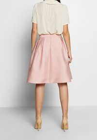 Falda acampanada de color rosa claro hecha de tejido suave, con pliegues, combinada con una blusa crema y unos tacones beige texturizados.