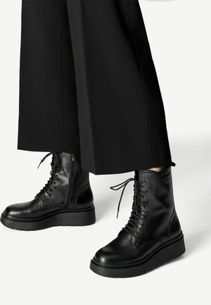 Black leather lace-up combat boots with side zipper and thick sole worn with wide-leg black pants over white background.