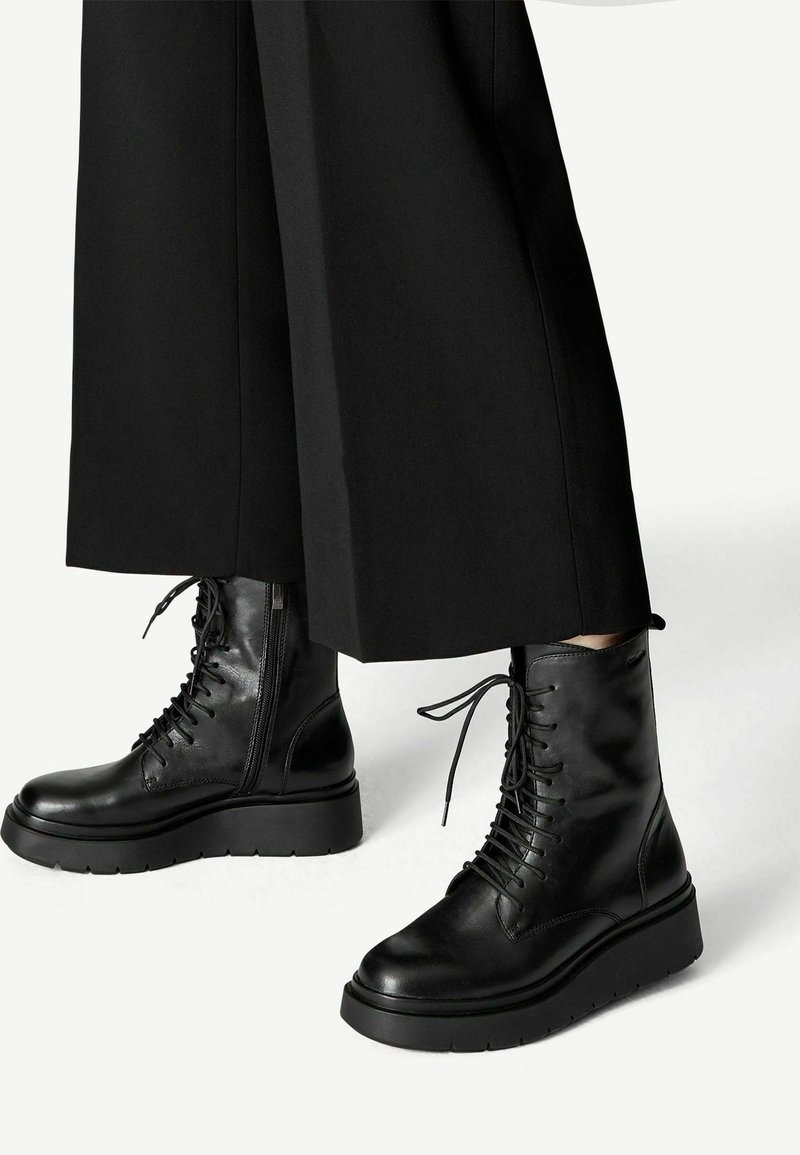 Bottes de combat montantes en cuir noir avec lacets, fermeture éclair latérale et semelle épaisse, portées avec un pantalon noir à jambes larges sur fond blanc.