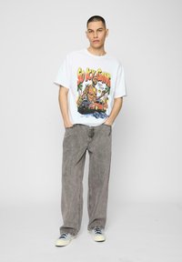 Hvid grafisk t-shirt med "So Icy Gang" design, parret med grå brede bukser og sneakers med blå accenter, på ensfarvet baggrund.