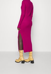 Robe en maille côtelée fuchsia à manches longues avec une fente latérale, associée à des bottines jaunes à talon bloc et texture brillante.