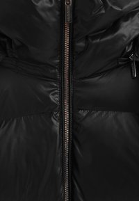 Svart vadderad jacka med central dragkedja, slät blank yta och subtila paneldetaljer. Dragkedjeförsedd med synlig varumärkeslogotyp.