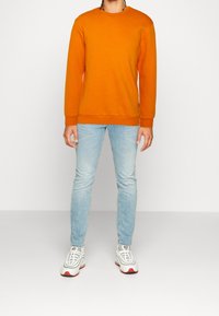 Felpa arancione, jeans azzurri, scarpe da ginnastica bianche con dettagli rossi; presenta scollo rotondo, maniche lunghe e vestibilità comoda.