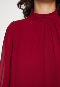 Blouse bordeaux avec un col haut, un design plissé et des manches longues transparentes, fabriquée en tissu léger. Texture lisse.