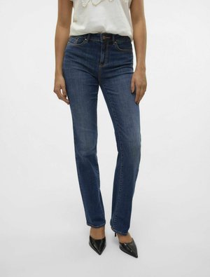 Jeans de denim azul de cintura alta con corte ajustado, que presentan áreas deslavadas, costuras en contraste y cierre de botón. Combinados con tacones negros.