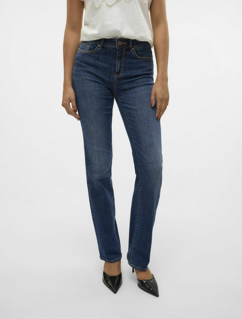Jeans de denim azul de cintura alta con corte ajustado, que presentan áreas deslavadas, costuras en contraste y cierre de botón. Combinados con tacones negros.