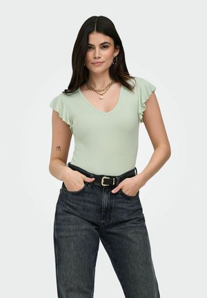 Woman wearing light green ruffled sleeve top, black high-waist jeans with belt, gold chain necklace, hoop earrings, hands in pockets.