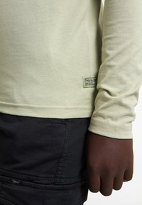 Lichtgroen shirt met lange mouwen en een zachte textuur, voorzien van een zwart stoffen label aan de onderkant, gecombineerd met een donkere short.