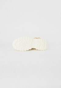 Sneaker blanc avec une semelle en caoutchouc texturée, présentant un design imposant et des lacets plats. La semelle a des rainures profondes pour la traction. Pas de marquage visible.