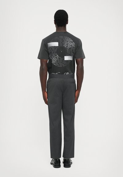 Man in dark graphic t-shirt with circular text design, gray trousers, and black shoes stands facing a white background.