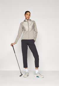 Giacca zip-up beige chiaro con un pannello del petto imbottito in argento, abbinata a pantaloni grigio scuro e scarpe da ginnastica beige. Tiene una mazza da golf.