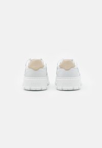 Weiße Sneakers mit dicker Sohle, Wildlederakzenten in Beige, gepolstertem Schaft und erhobener Markenkennzeichnung an der Ferse. Glatte Textur rundum.